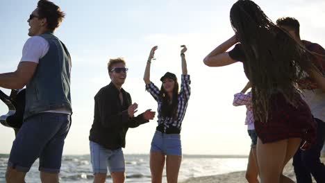 summer party on the beach. young friends drinking cocktails, dancing in the cirkle, playing guitar, singing songs and clapping on a beach at the water's edge during the sunset. slowmotion shot