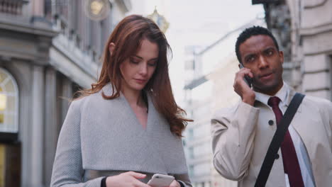 Millennial-white-woman-standing-on-the-street-using-smartphone,-young-black-businessman-talking-on-phone-walking-past,-close-up,-low-angle