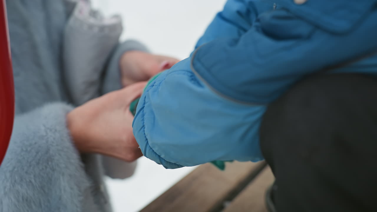 Closeup hands putting sock on son, winter bench scene mom gently pulls striped knitted sock onto childs foot, blue coat and wooden bench, snow in background, warm caring interaction and intimate