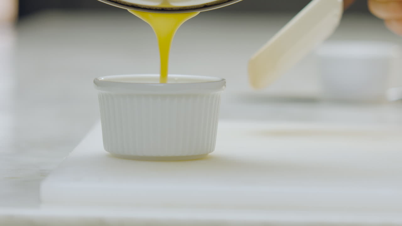 Close-up of a chef pouring creme brulee from a strainer directly into a white ceramic mold on a chopping board