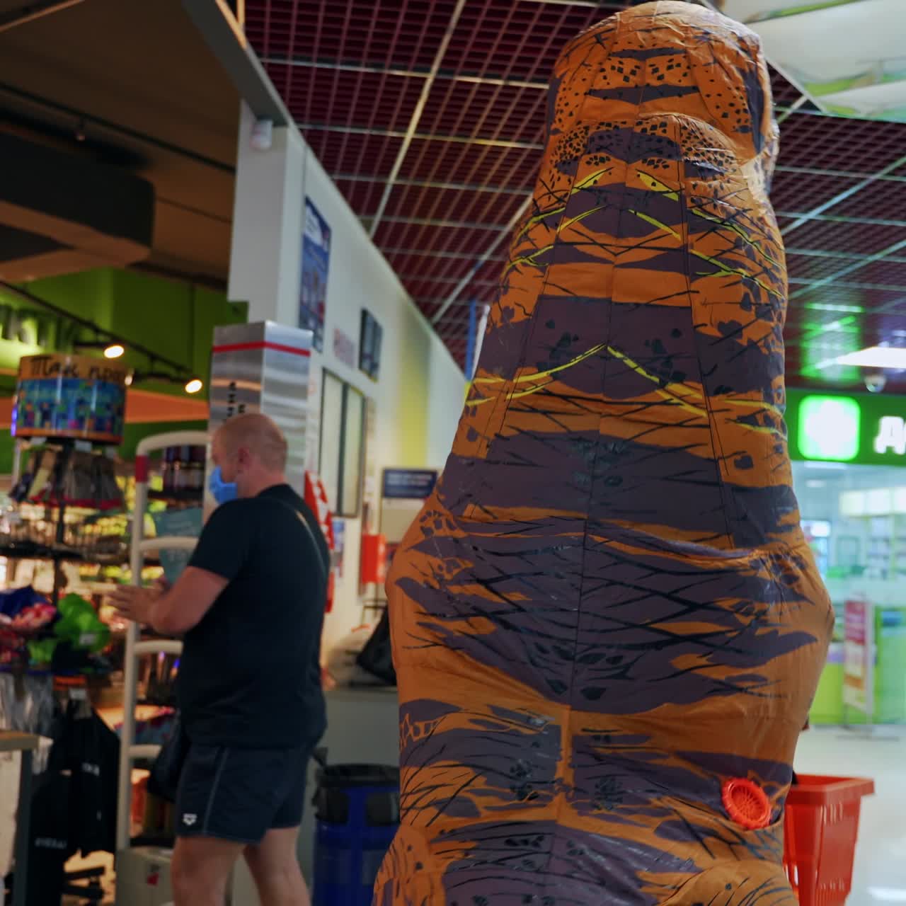 Animator in orange dinosaur costume walking by the supermarket. Man in funny inflatable costume holding a shopping basket