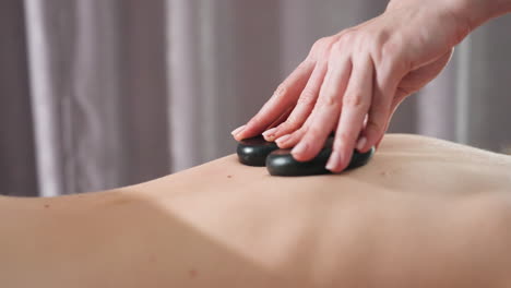 close view of massage therapist's hands using two smooth black stones to massage client s bare back in soft-lit spa setting with gray curtain in background, focusing on technique and calming therapy