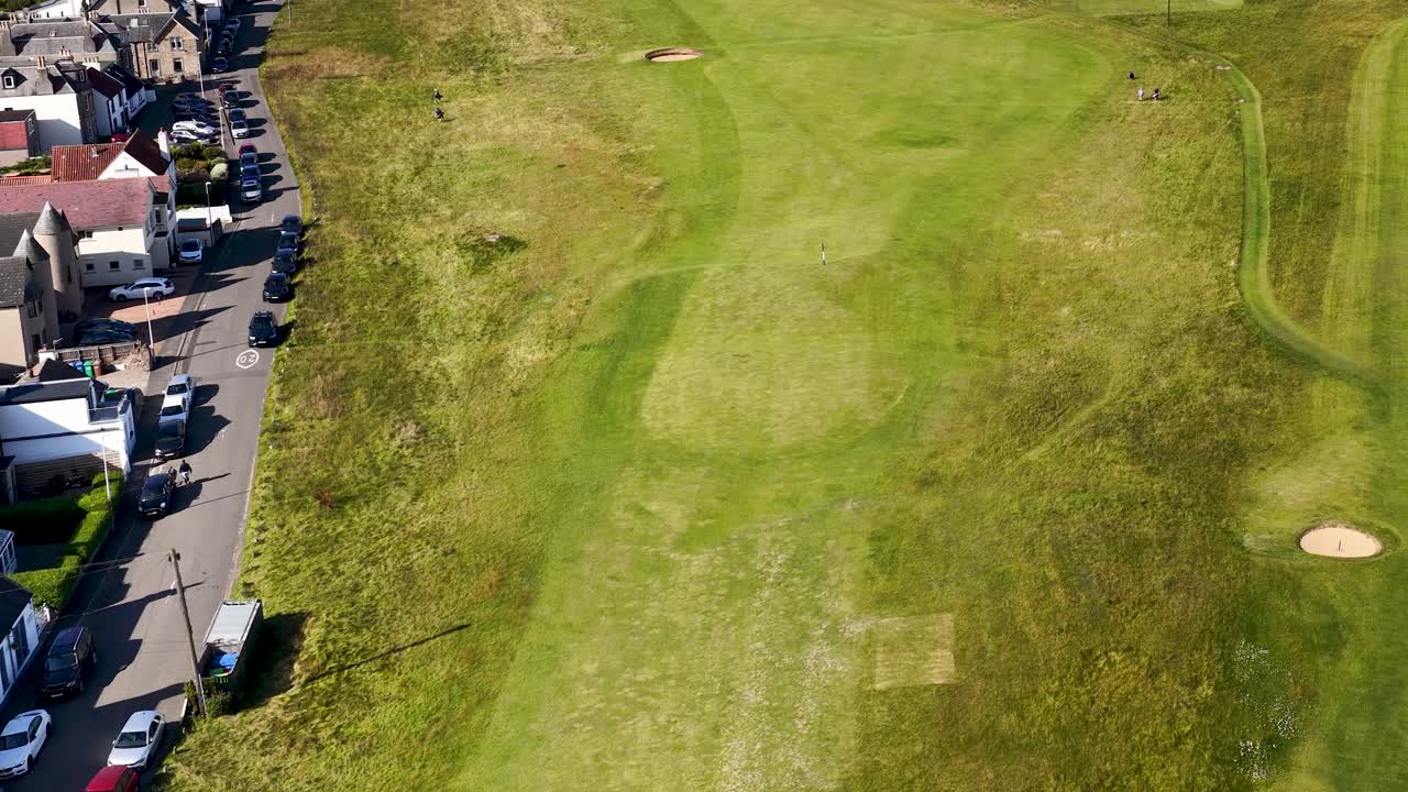 Drone glides above green golf course and village, bright daylight, smooth forward aerial movement