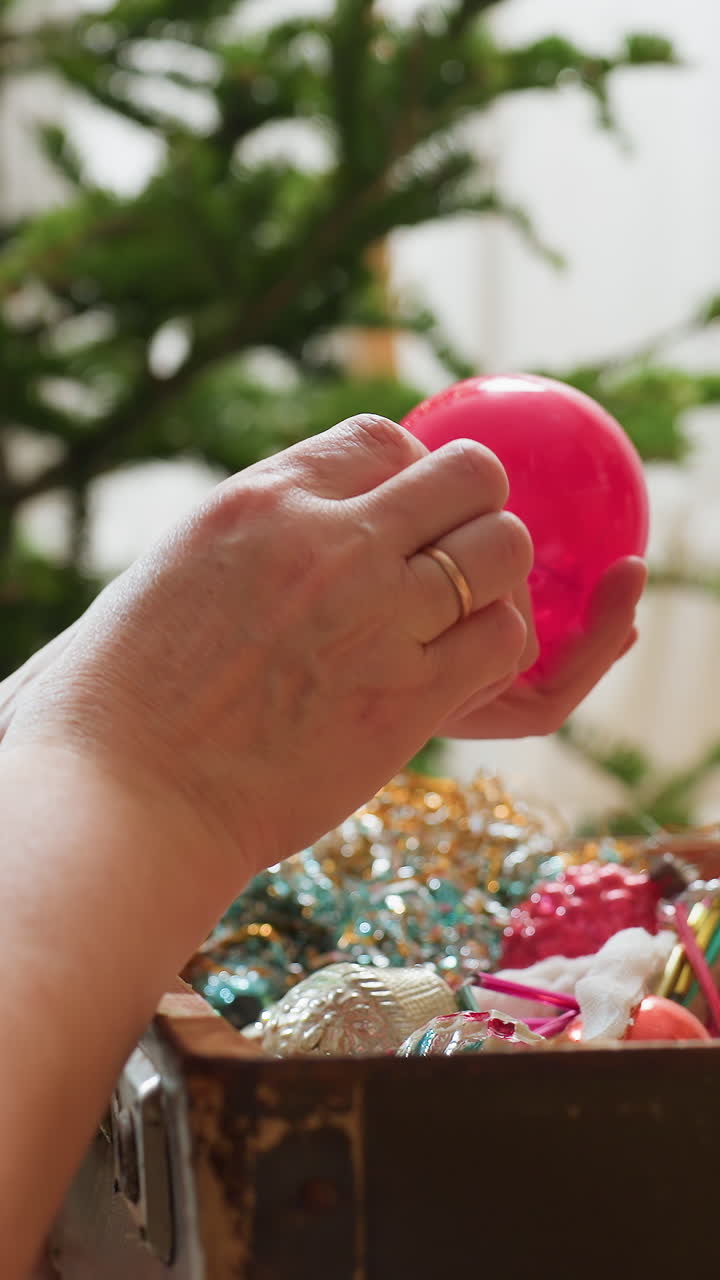 다채로운 장신구를 다루는 백인 여성 클로즈업 사진입니다. 반짝이는 장식, 분홍색과 보라색 유리 장신구를 소중히 다루는 손, 부드러운 트리 조명이 배경에서 흐릿하게 빛나고 있으며, 섬세하게 여행 가방 안에 넣고 있습니다.