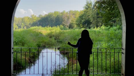 Silhouette-of-a-young-woman-spinning-through-the-frame-in-front-of-an-archway,-with-beautiful-lush-foliage-and-a-creek-in-background