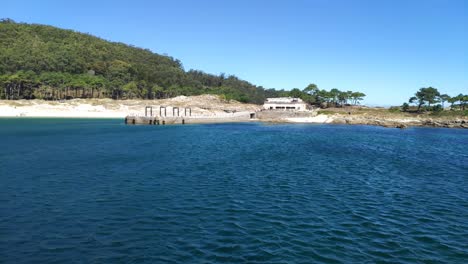 Jetty-and-restaurant-in-the-CÃ­es-Islands-with-the-beach-and-the-forest-of-the-natural-park-in-the-background-a-sunny-day-of-blue-sky,-blocked-shot-traveling-forward,-Pontevedra,-Galicia,-Spain