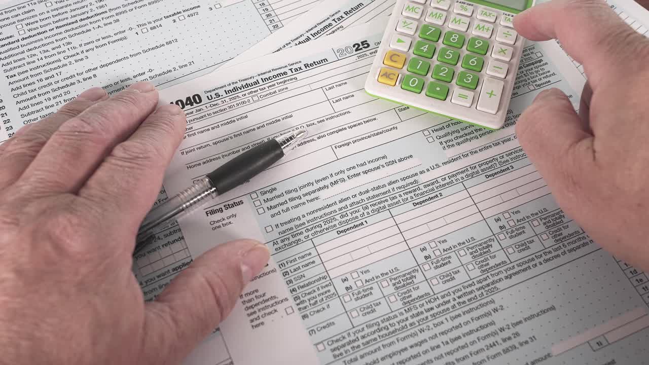 Hands are seen working on U.S. income tax 1040 forms while using a calculator. A pen is nearby for writing. The setting is a simple desk at home