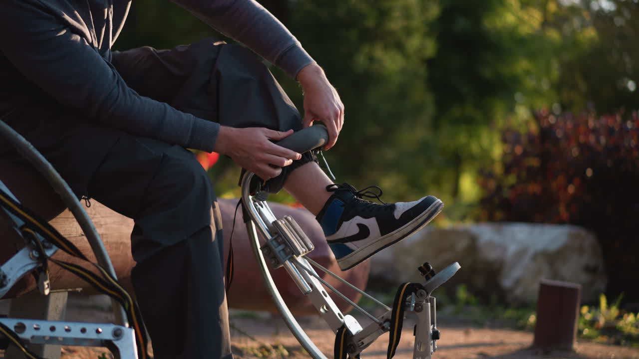 hombre sentado al aire libre con sudadera gris con capucha y pantalones oscuros, centrado en usar zancos con vegetación en el fondo. zapatillas de deporte negras y blancas son visibles, añadiendo un toque casual al aire libre centrado