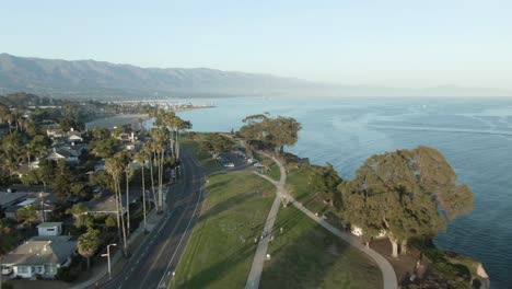 High-altitude-aerial-over-coastline-city-park-during-sunset-with-trees-and-the-Pacific-ocean-with-mountains-in-the-background,-in-Santa-Barbara,-California,-USA