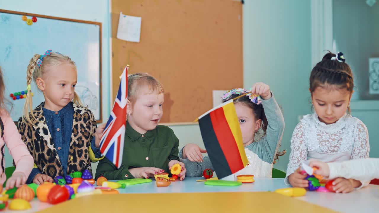 Children learning English at the table with layouts of fruits in class. Two little girls holding flags and waving them on the background of education center. British and German flags.