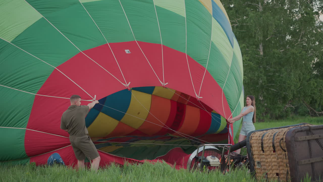 couple hold hot air balloon envelope open while burner fan heats air into vibrant checker patterned fabric for inflation during calm sunset pre flight preparation in serene grassy field