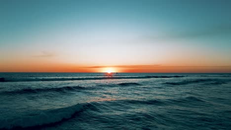 Flight-over-the-waves-of-the-Pacific-Ocean-towards-the-sunset-and-dusk-with-yacht-sailing-on-the-horizon