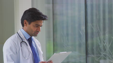 Male-Doctor-Wearing-White-Coat-Standing-In-Hospital-Corridor-Looking-At-Clipboard