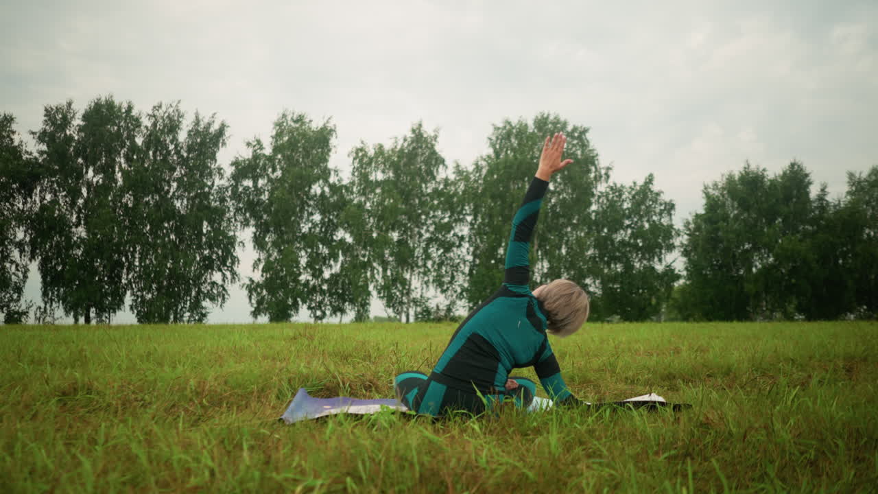 요가 매트의 한쪽에 누워있는 여성의 뒷면, 팔을 펴고 측면 구부러진 자세를 연습하고, 먼 곳의 자연과 나무로 둘러싸인 구름이 많은 하늘 아래 광활한 잔디에 앉아 있습니다.