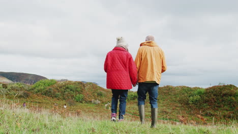 rear view of active senior couple holding hands as they walk along coastal path in fall