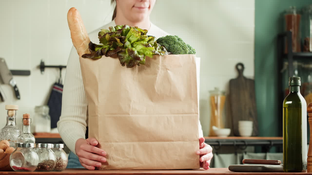 Woman holding a paper grocery bag filled with vegetables and bread in a kitchen
