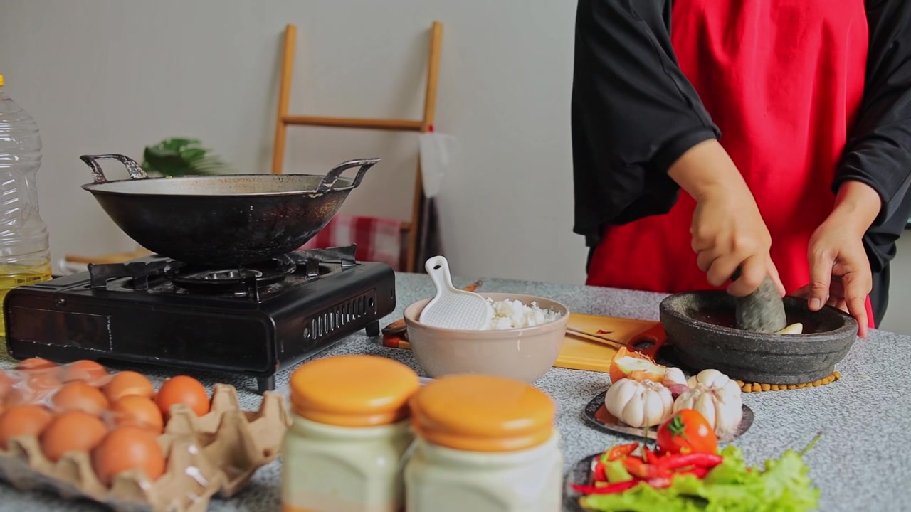 Tangan koki bercelemek menyiapkan bahan-bahan dan menghancurkan bawang putih menggunakan cobek batu dan ulekan di dapur