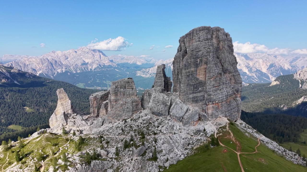 Veduta aerea con drone delle iconiche torri di calcare delle Cinque Torri, circondate da terreno roccioso e sentieri alpini, con ampie cime dolomitiche e valli boscose sullo sfondo