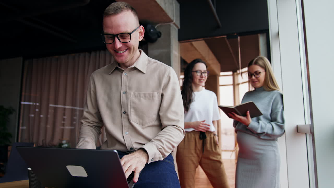 Happy laughing male in glasses holding laptop on his knees. Two women stand at backdrop talking. Pregnant blonde lady looks at paper book.