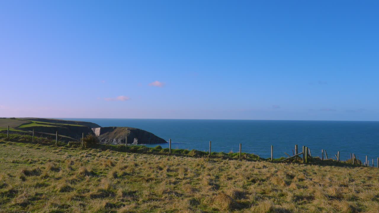Wide Angle Pan of Empty Farm Field on Welsh Coastline with Fence and Rocky Coast with Bright Blue Sky and Sun Flare.
