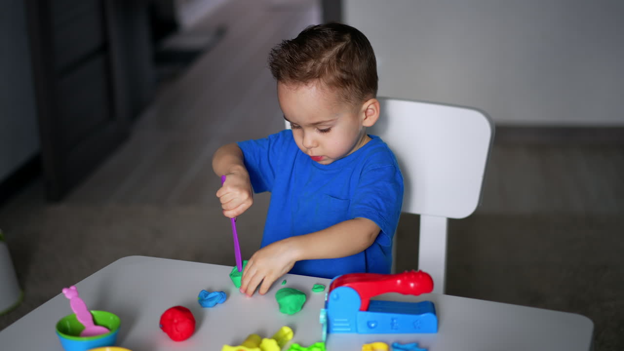Small kid sits at desk busy with his hobby. Cute toddler plays with plasticine and stick. Top view.