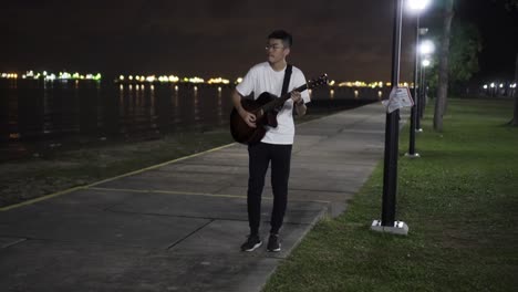 man playing guitar alone at night in east coast park, singapore while it is dark with the ships light and street light