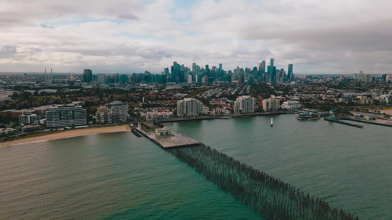 Filmato cinematografico 4K con drone che riprende Princes Pier, case costiere, il CBD di Melbourne all'orizzonte lontano e acque blu dell'oceano sotto cieli mattutini soleggiati e ventosi a Melbourne.