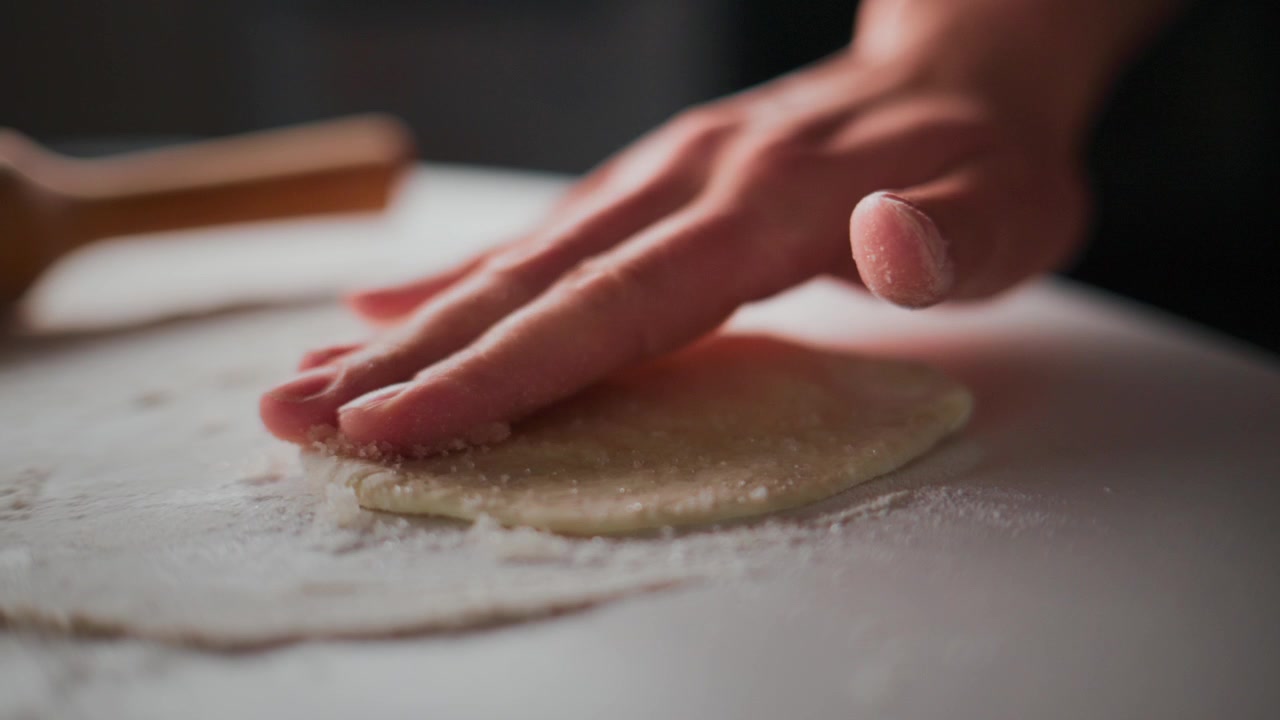 Una mano preme delicatamente l'impasto su una superficie infarinata. Le dita spargono la farina mentre l'impasto si appiattisce. Atmosfera calda e rustica da cucina. Ideale per ricette, tutorial di pasticceria o contenuti a tema culinario.