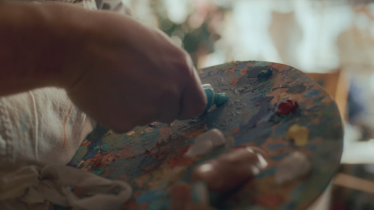 Primer plano de las manos de un artista exprimiendo pintura al óleo azul sobre una paleta de madera.