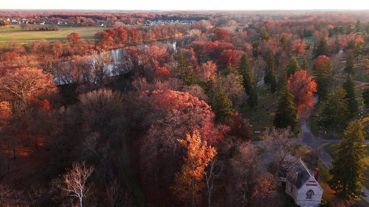 Drone view panning left over autumn trees next to river with church and cemetery