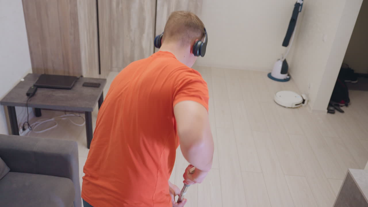 Vista posterior de un joven con una camiseta naranja y auriculares que sostiene una fregona como si fuera una guitarra mientras baila durante su rutina de limpieza en un salón luminoso, expresando alegría