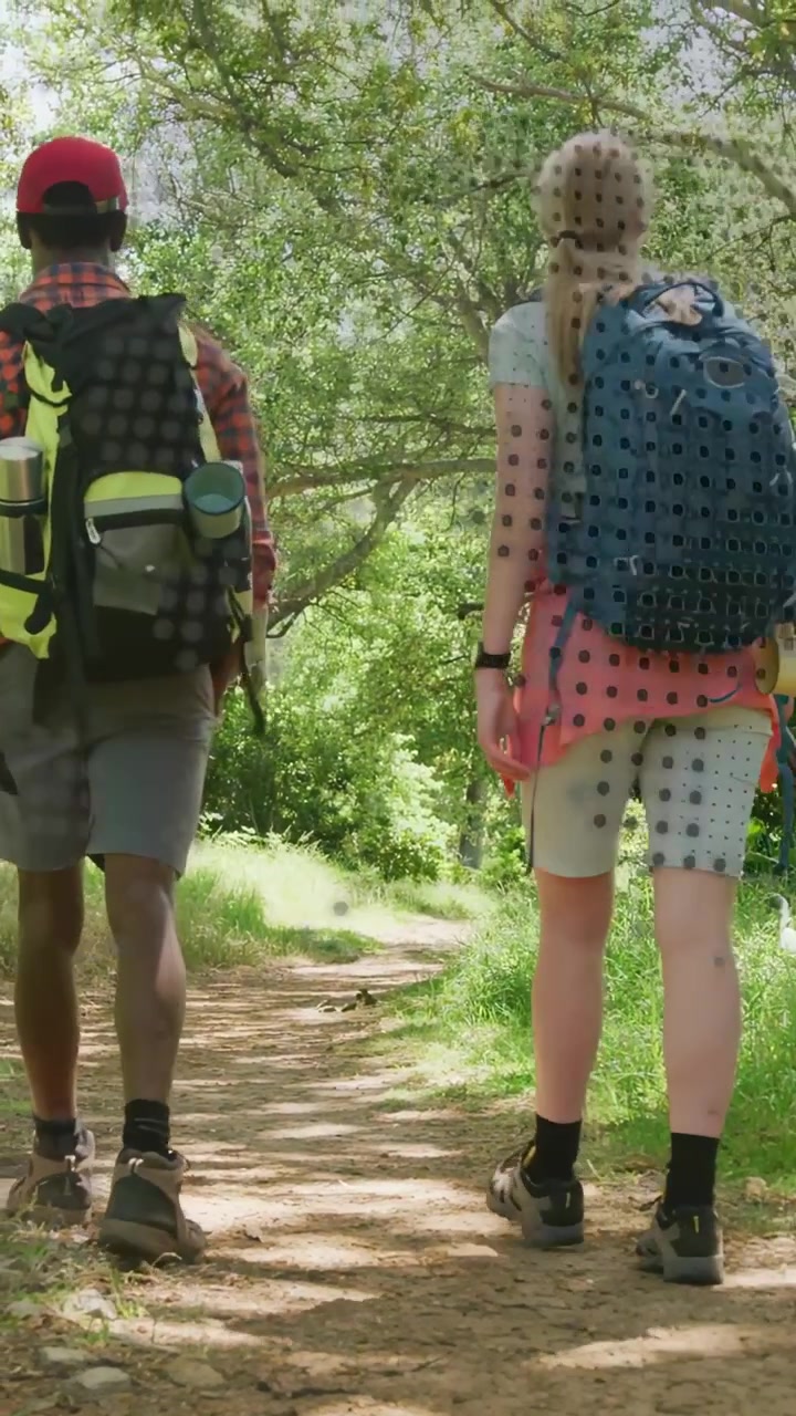 Video verticale: Due escursionisti all'aperto che camminano su un sentiero, la telecamera riprende un cappellino rosso e uno zaino blu verso una radura