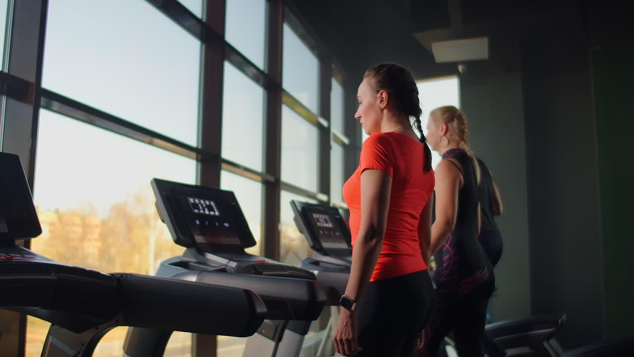 A group of people walking on treadmills near a large panoramic window. Group cardio workout. Start of fitness classes. Young beautiful women and men in sportswear in cardio fitness room.