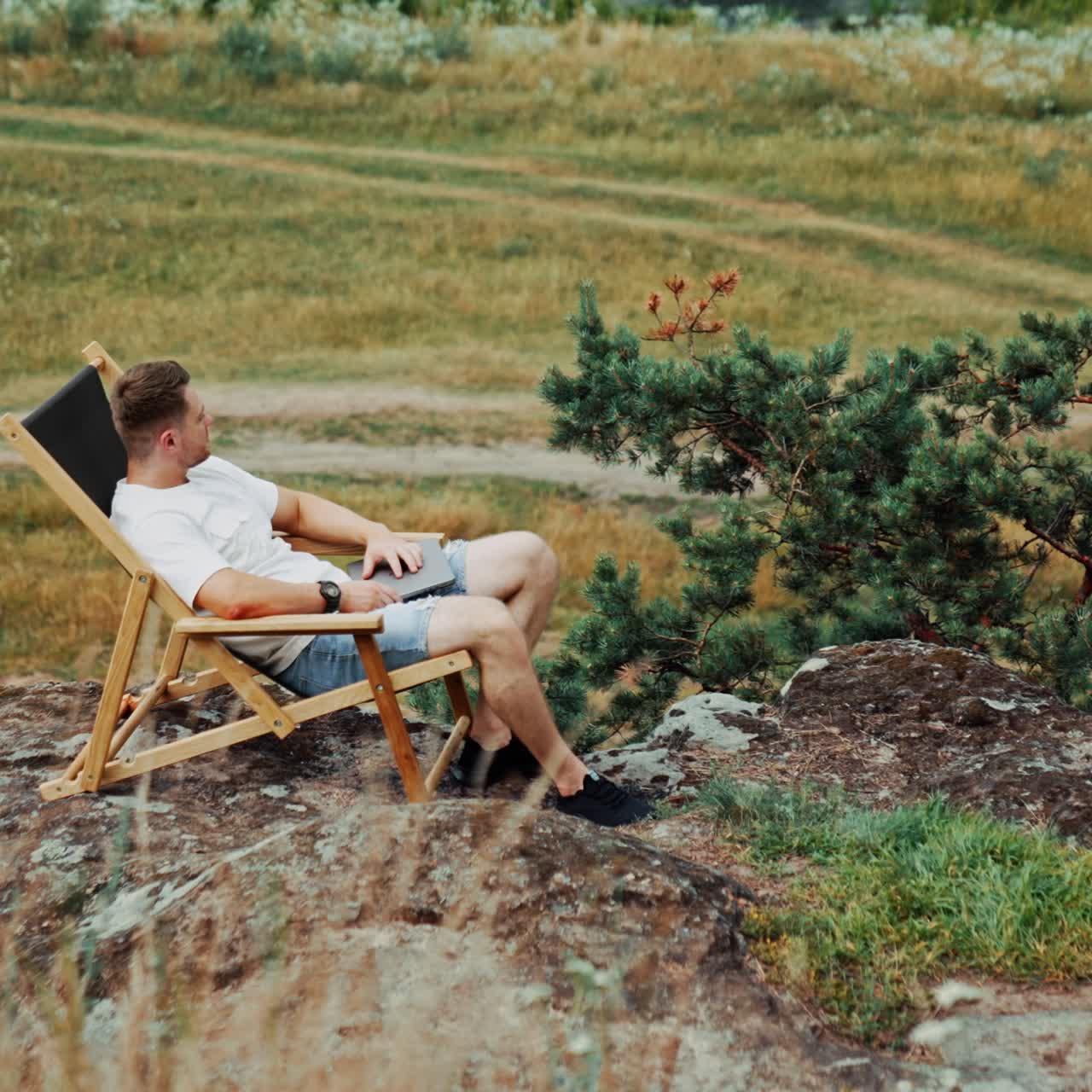 Pensive relaxed male sits in a folding chair on the rock. Man looks around enjoying the view after work on laptop. High angle view