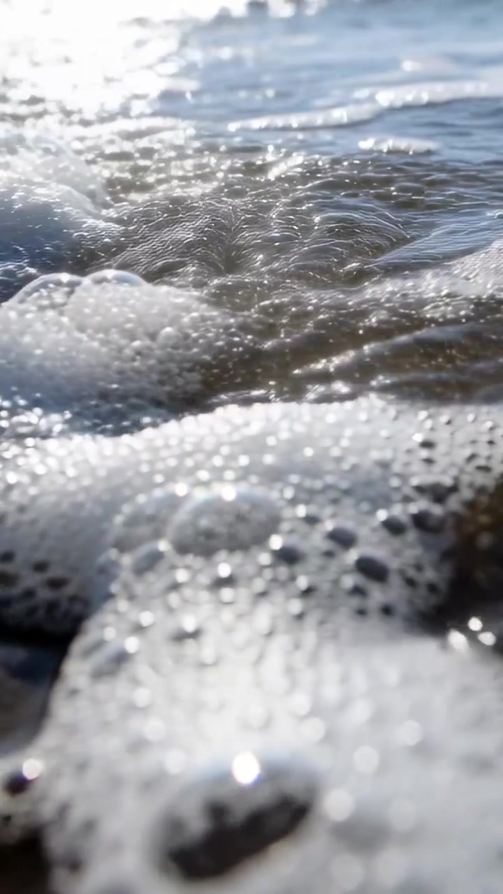 Video verticale: Schiuma che si diffonde, bolle che scorrono sulla riva di sabbia bagnata, un'onda che spinge il riflesso scintillante del sole.