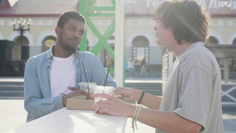 male friends chatting while sharing pizza and drinking, standing at an outdoor table in the street