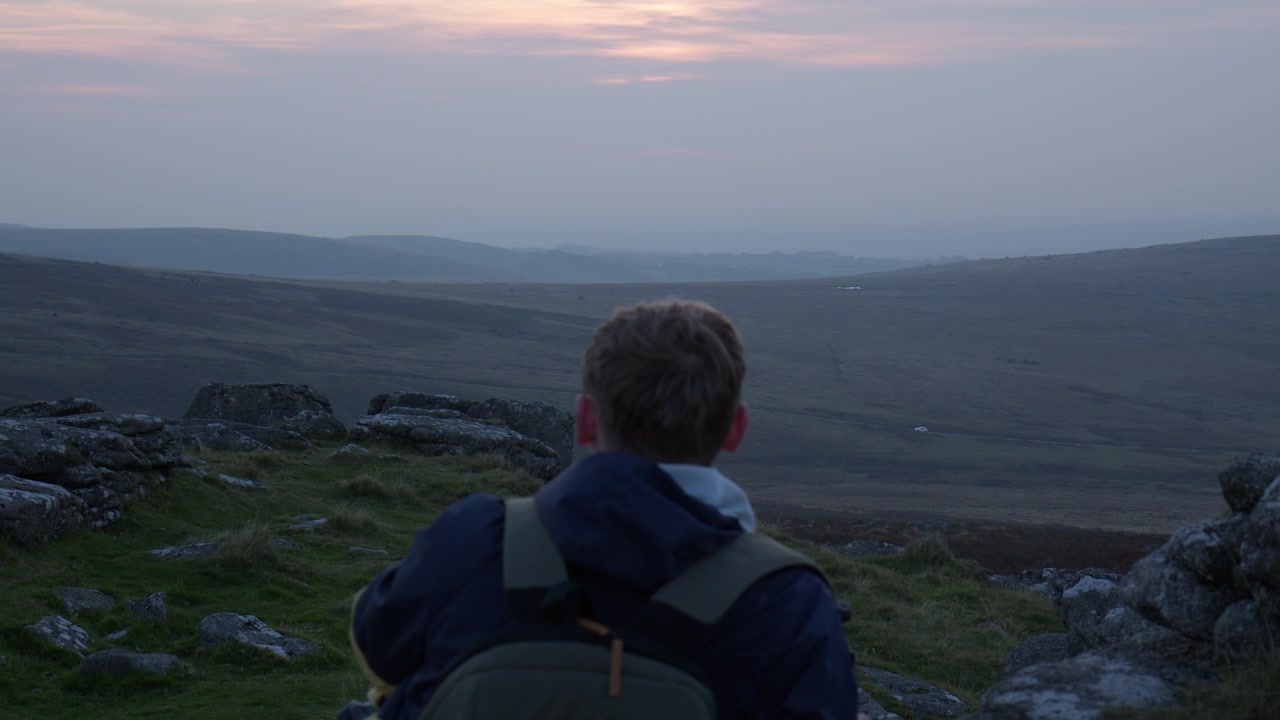 Dartmoor, Devon, Inghilterra, Regno Unito. Una persona con uno zaino siede su un fianco di collina roccioso, ammirando colline ondulate sotto un cielo al tramonto fioco. Primo piano.
