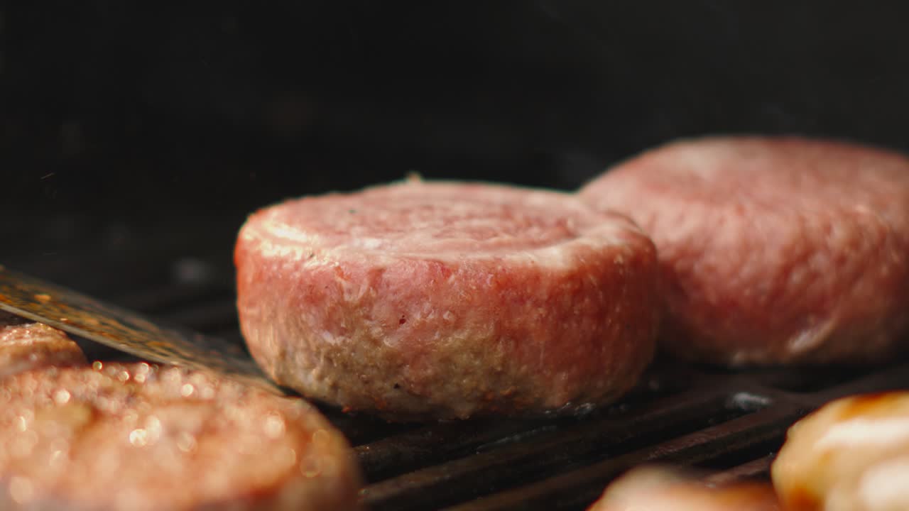 Close Up Flipping Large Raw Beef Burger on Simmering BBQ Grill with Other Patties Cooking. Delicious Summer Food Being Cooked Outdoors