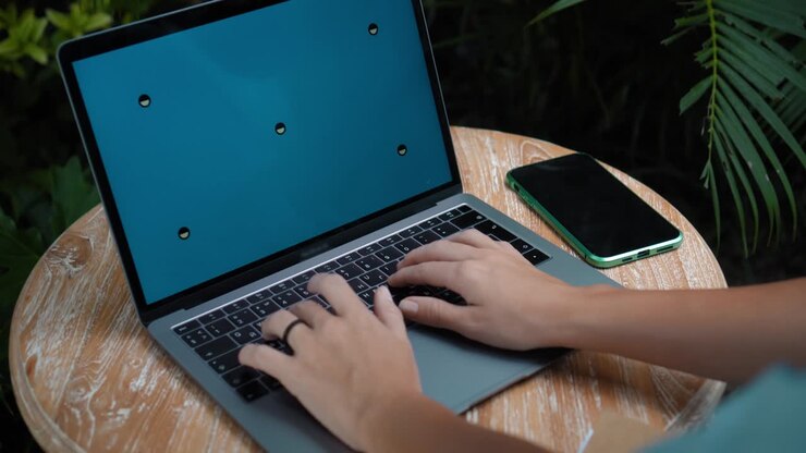 Eine junge Europäerin in einem blauen Kleid arbeitet remote online an einem Laptop mit blauem Bildschirm im Garten mit grünen Pflanzen