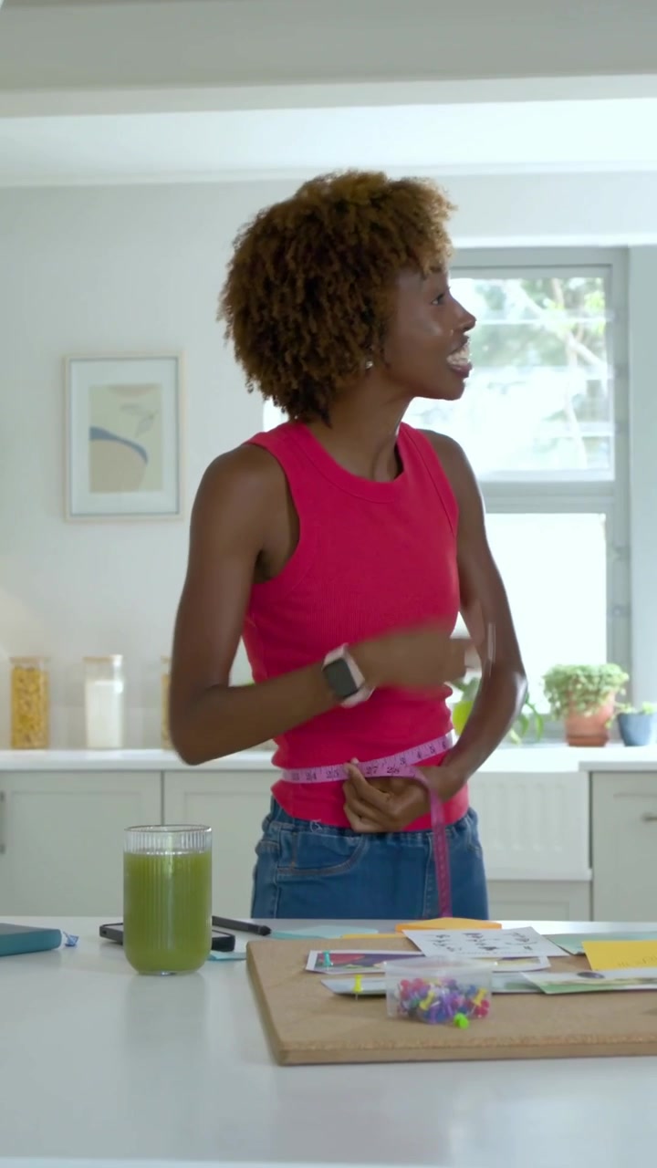 Video verticale: Donna con un top rosa che raccoglie, misura e lega un nastro in cucina, provando la vestibilità.