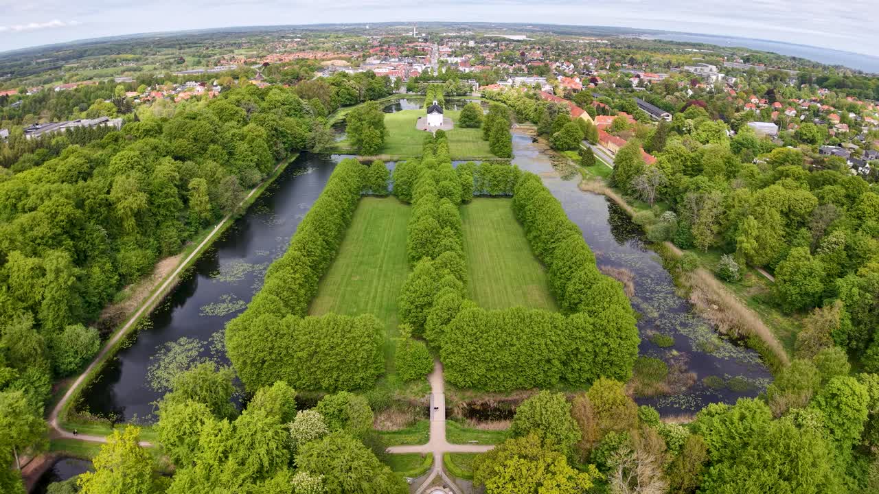Drone footage offers a top-down view of a long, tree-lined canal or park in Denmark. Symmetrical rows of green trees border the narrow waterway, creating a striking geometric pattern in the landscape