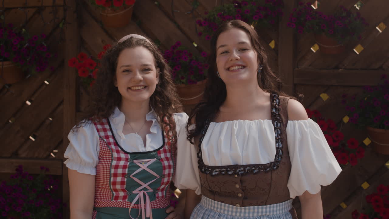 Two women in traditional Bavarian dresses smiling in front of a flower-decorated background