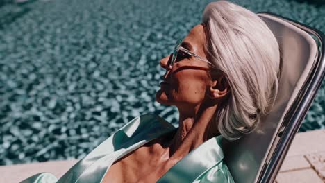 elegant senior woman enjoying a moment of tranquility by the pool, wearing sunglasses and a light green satin robe, embodying a sense of sophisticated relaxation and timeless beauty