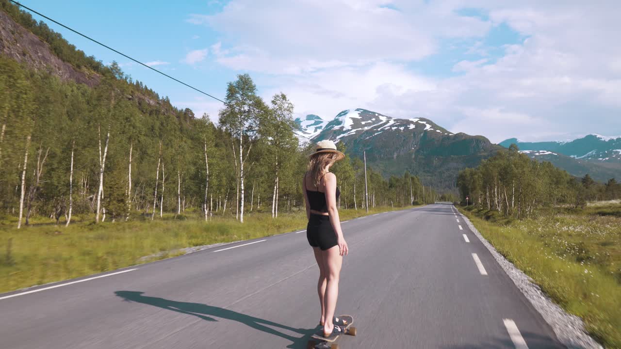 Young girl skateboarding down a large street without cars, shot taken from behind, girl looking towards the camera. Mountain and forest surroundings in Norway.