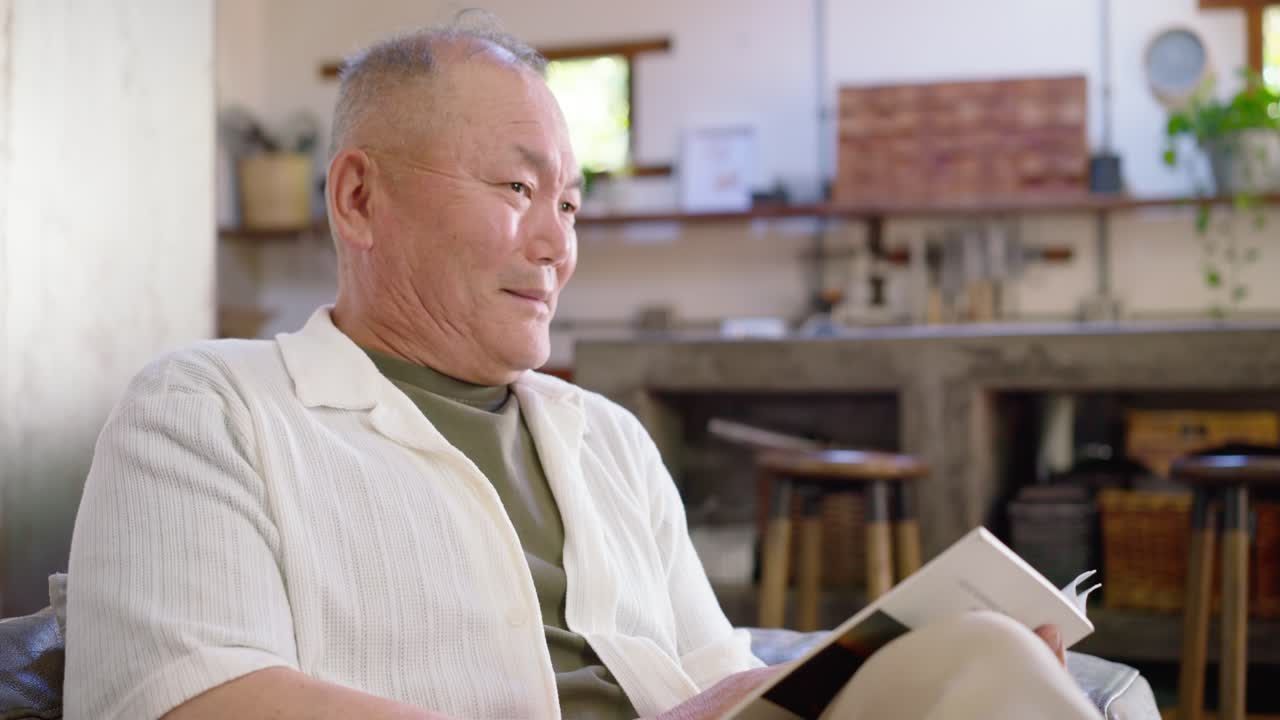 ソファでペーパーバックを読むアジア系の高齢男性。窓際でページをめくりながら、その一節に反応している。