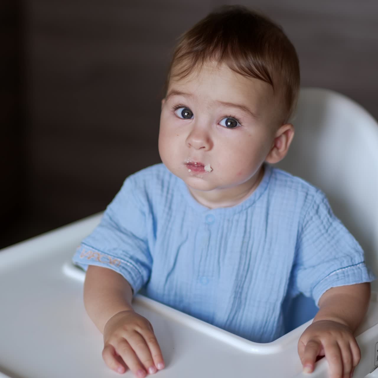 Sweet kid in blue shirt sits at feeding table. Mother giving a full spoon to her child. Close up. Blurred backdrop