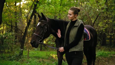 joven feliz jockey hembra está acariciando a su caballo marrón con mancha blanca en la frente en el parque durante un día soleado sosteniendo correa de cuero