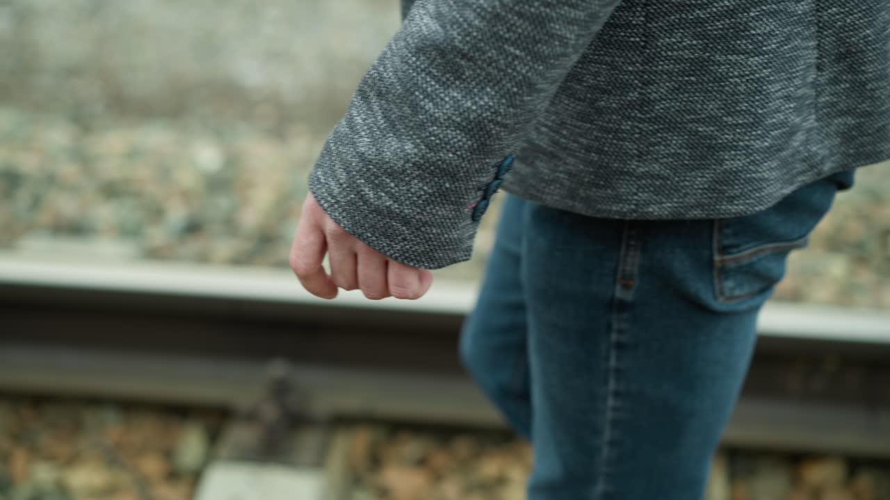 회색 슈트 재과 파란 청바지를 입은 남자의 손이 철도 트랙을 따라 고있는 근접 시각, 그의 손은 에 편안하고, 배경에는 흐릿한 철도 돌과 트랙이 표시됩니다.