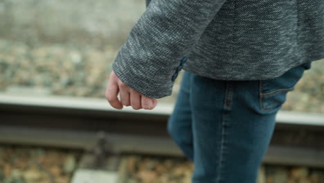 a close-up view of a man's hand wearing a grey suit jacket and blue jeans, walking along railway tracks, his hand is relaxed by his side, with the background showing blurred railway stones and tracks