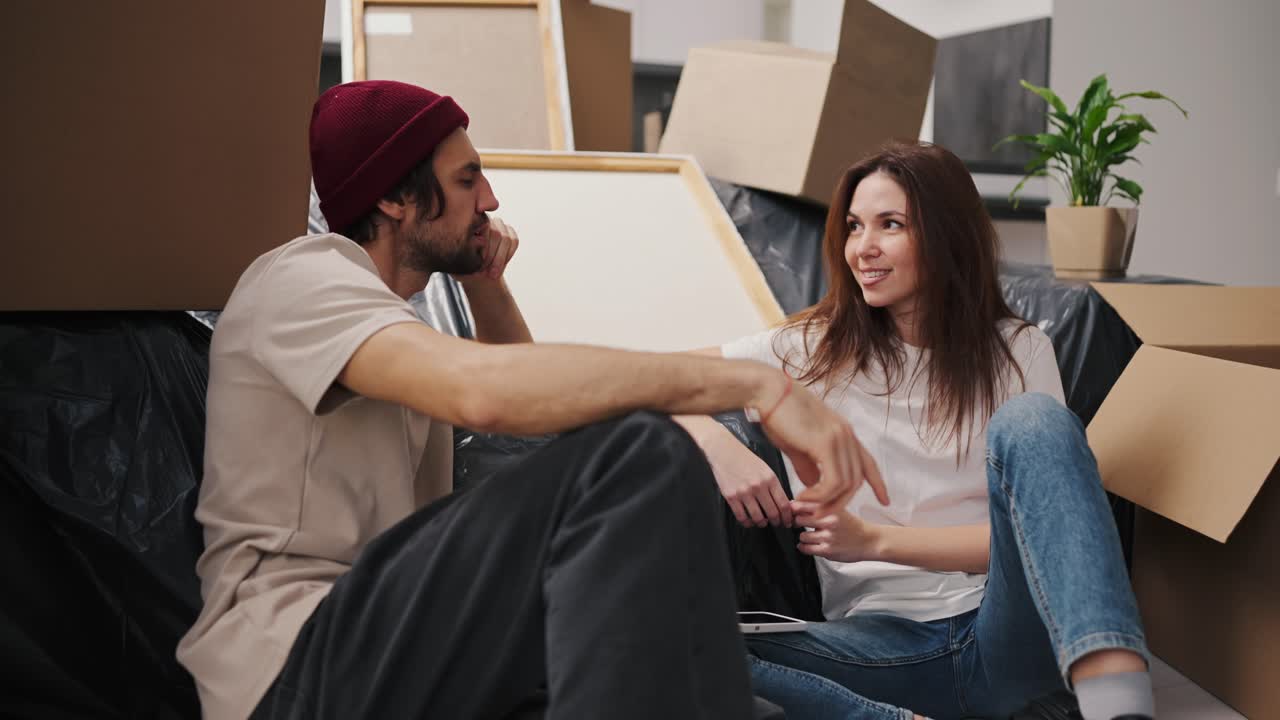 una chica morena con una camiseta blanca se comunica con su novio en un sombrero rojo con paja mientras se sientan cerca de un sofá cubierto de polietileno negro entre un gran número de cajas y una planta de la casa en un moderno